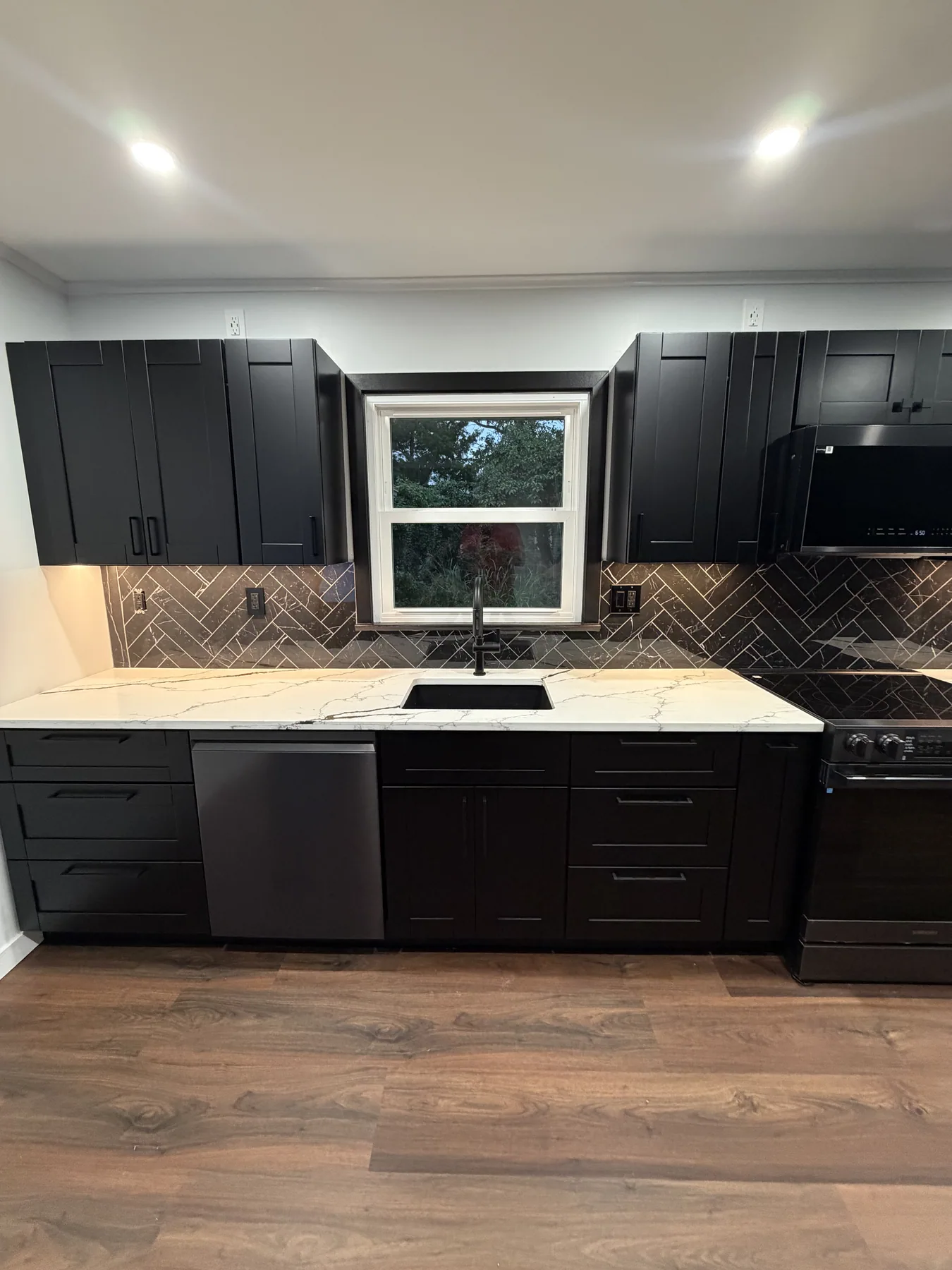 Dark kitchen cabinets framing a window above a black sink and light countertop