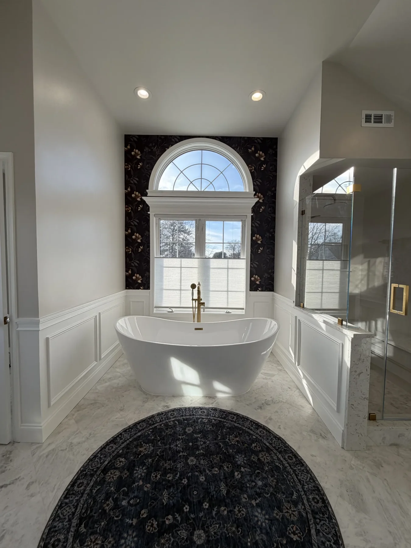 White freestanding bathtub beneath an arched window, with brass fixtures, white wall paneling and a glass shower